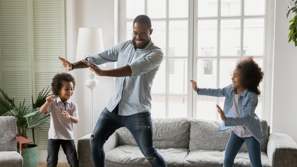 A Father and two young children on the left and right of the dad are all dancing in a room with a chair couch window and lamp behind them