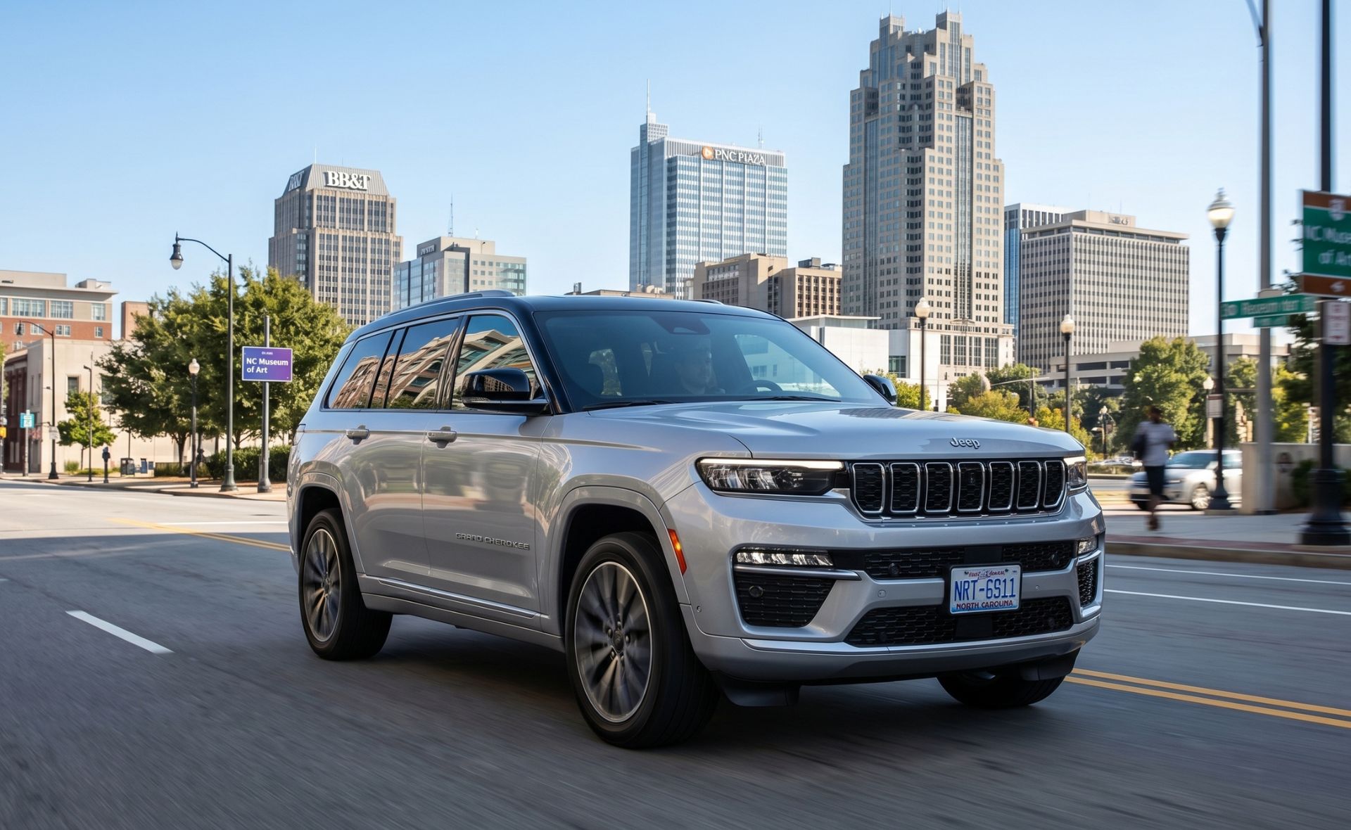 A silver Jeep Grand Cherokee luxury SUV driving down a city street in downtown Raleigh, featuring the iconic PNC Plaza skyscraper and urban skyline in the background.