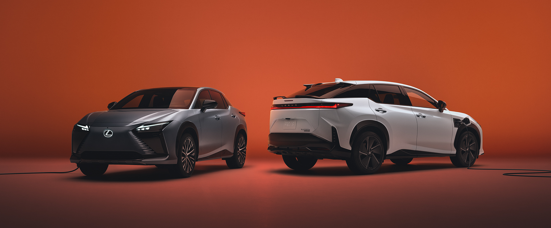 Two Lexus RZ electric SUVs, silver and white, plugged into chargers against a warm orange gradient background.