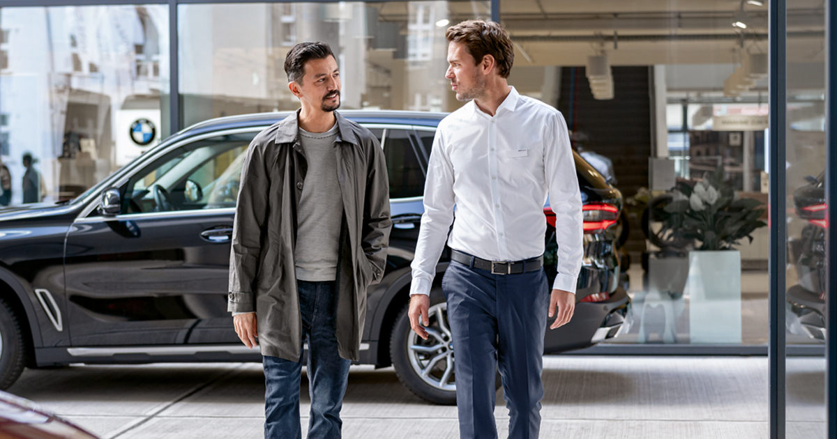 Two men walking together outside a modern building near a dark-colored BMW vehicle.