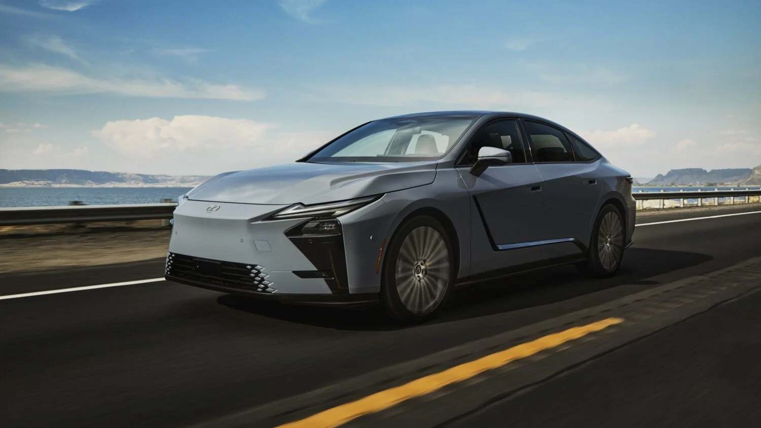A gray Lexus sedan driving along a coastal highway beside a guardrail, with water and bluffs visible in the background.