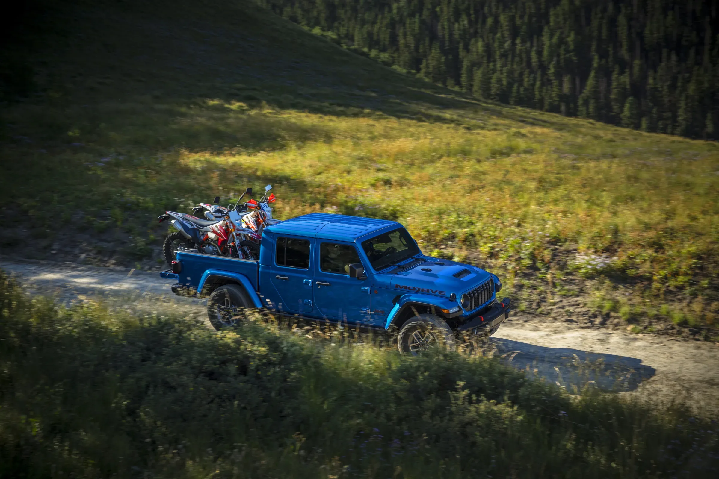 Blue Jeep Mojave-trim pickup truck carrying two dirt bikes on a mountain trail