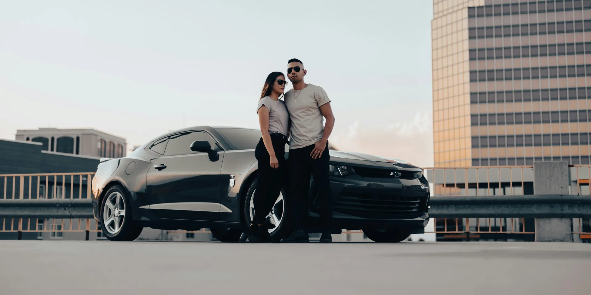 Man and woman in sunglasses standing close together beside a black Chevrolet Camaro on a rooftop parking level, city skyline in the background at dusk
