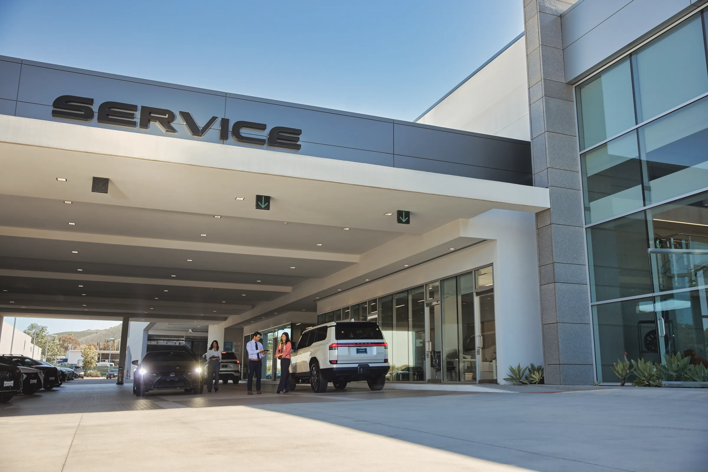 Modern Lexus dealership SERVICE canopy with a white Lexus SUV in the service lane while an advisor with a tablet greets customers for complimentary maintenance.