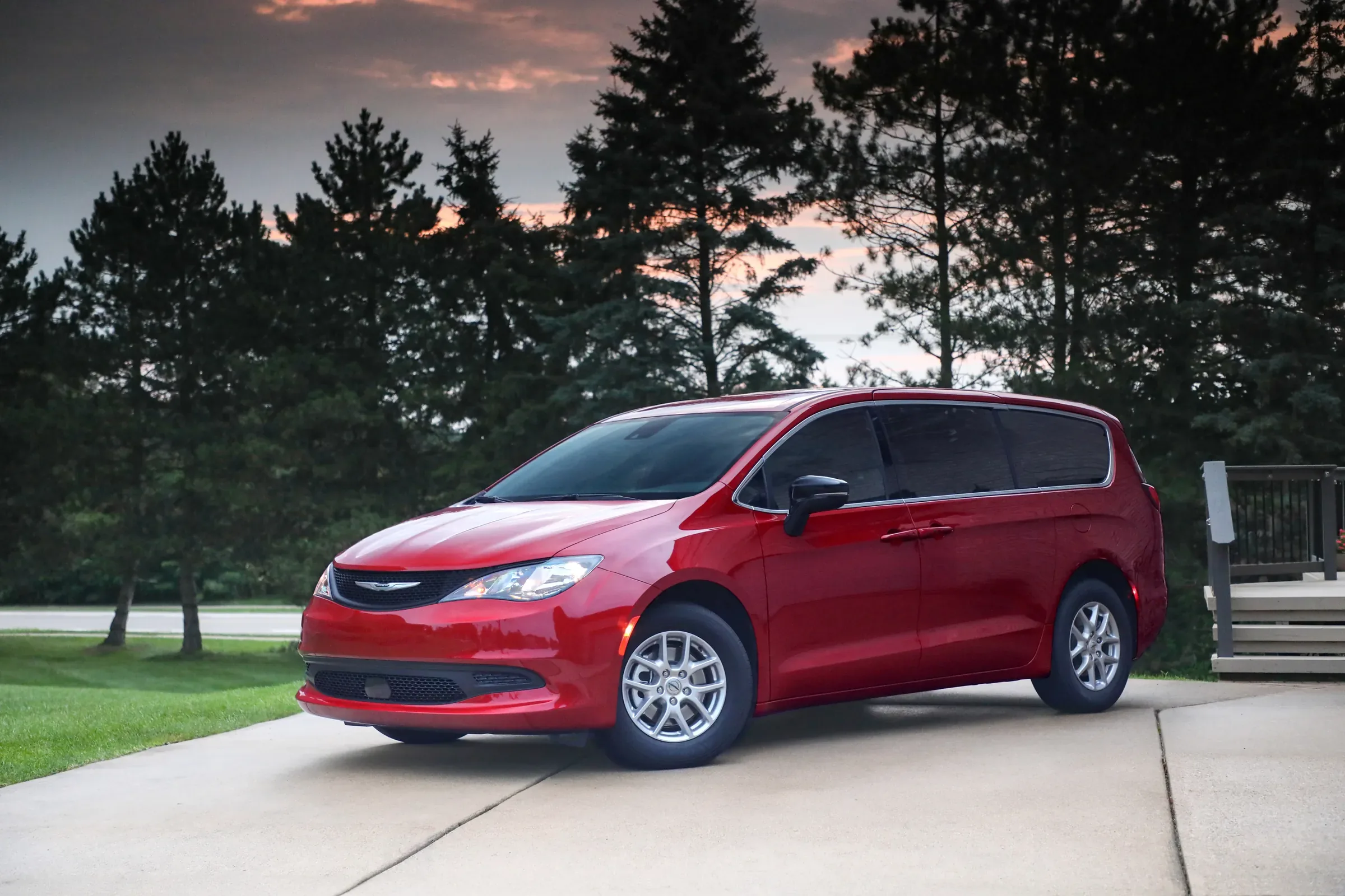 2026 Red Chrysler minivan parked in a residential driveway at dusk, backed by a row of tall pine trees