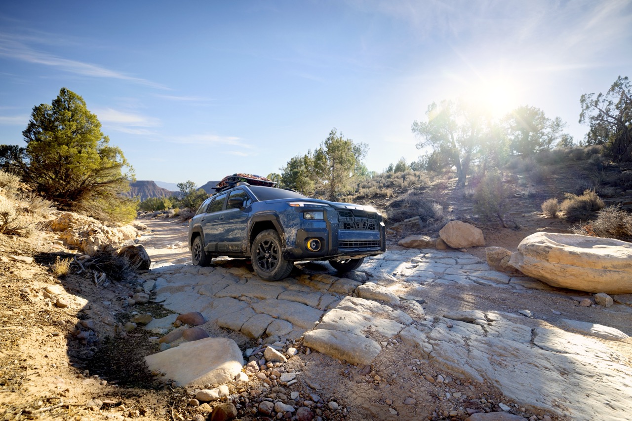 A static image of a blue 2026 Subaru Outback Wilderness driving slowly over an uneven rocky trail in a dry desert-like landscape. 