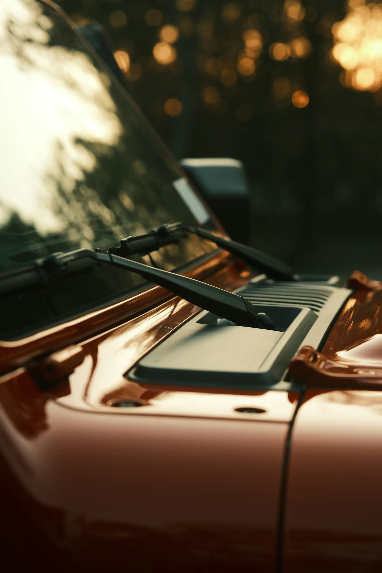 Close-up of an orange SUV's hood, windshield wipers, and cowl vents backlit by a sunset. Windshield Wiper Service Near Kansas City