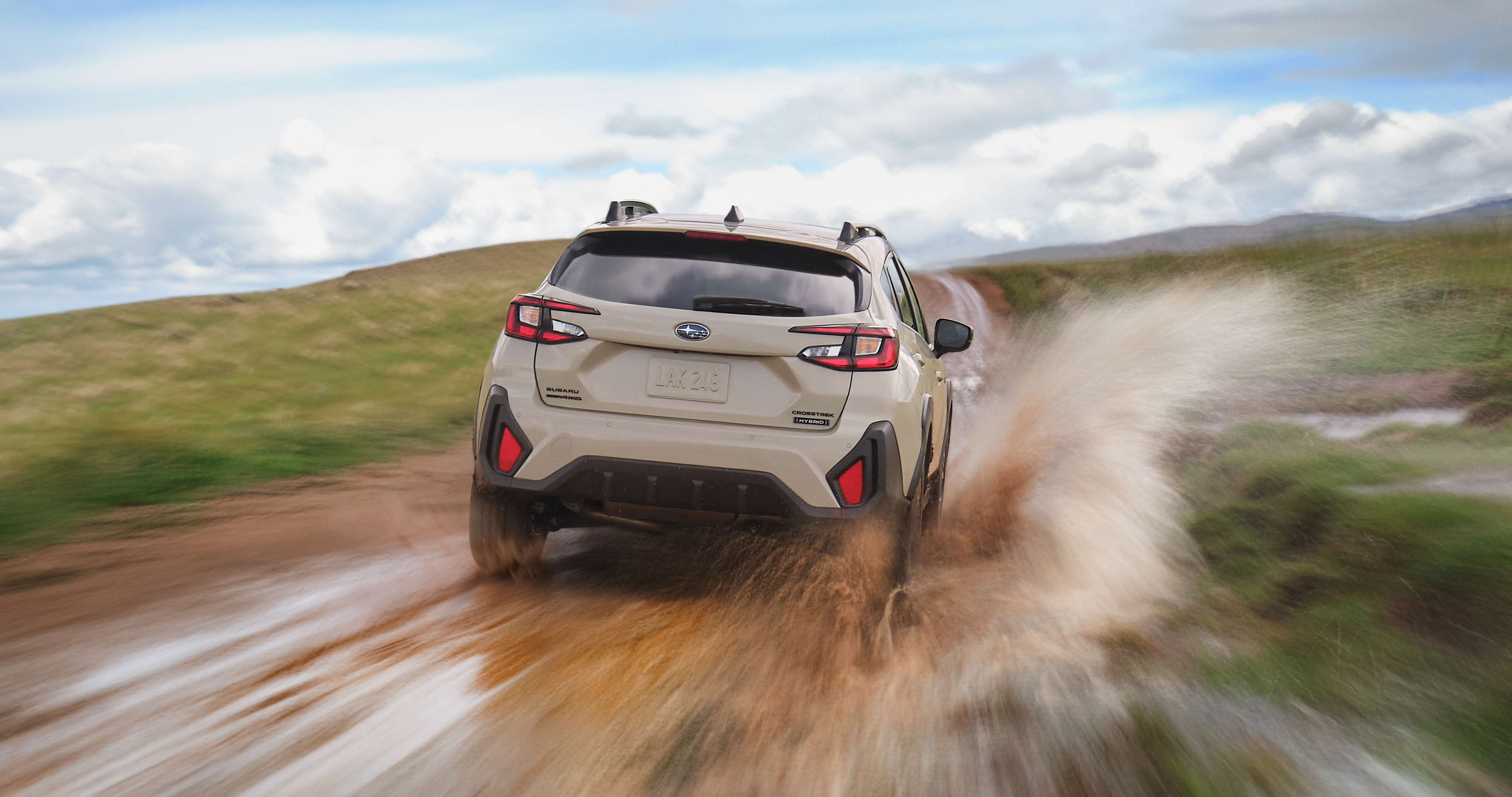 A Subaru Crosstrek Hybrid driving at speed down a muddy trail, kicking up water and mud as it moves through an open landscape under a partly cloudy sky.