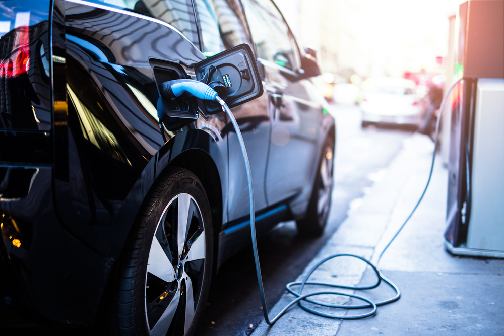 EV car is being charged by a Teal EV charger with a Teal cord, the car being charged is black, in the background you see a silver car up ahead