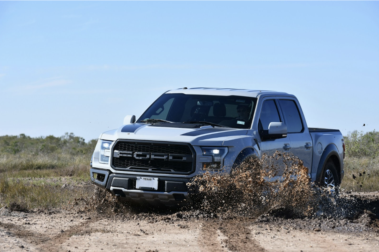 A gray Ford truck splashes through mud and water on a dirt trail, with brush and a clear blue sky in the background.
