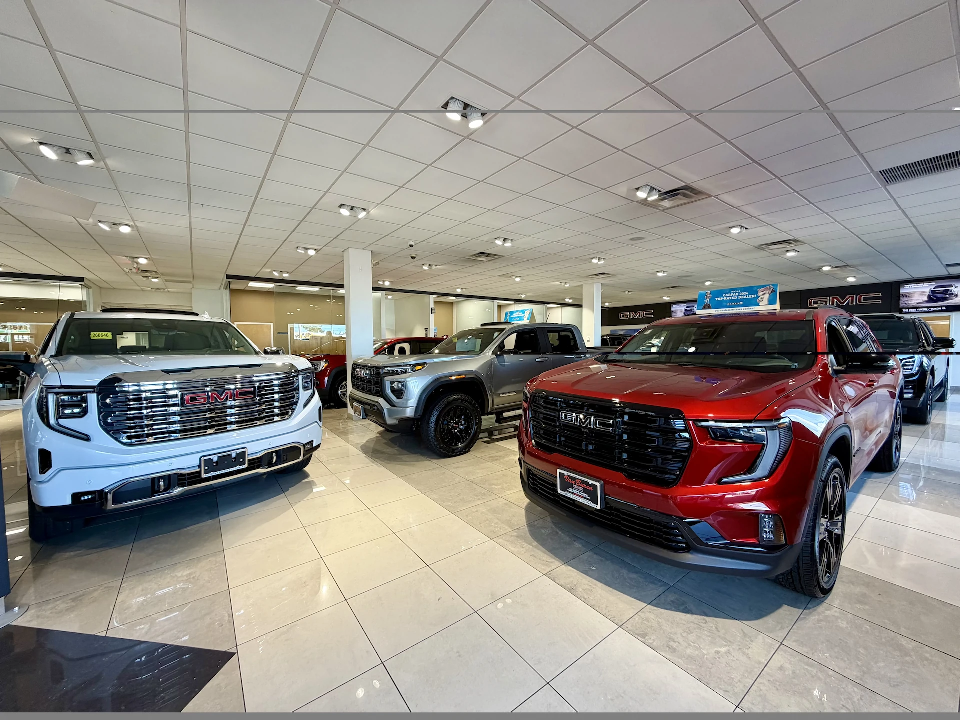 A white SUV, a silver off-road-styled pickup truck, and a red SUV displayed side by side in the Van Buren GMC showroom, each with a legible GMC grille badge, under a tiled ceiling with additional vehicles and a Carfax Top-Rated Dealer sign visible in the background