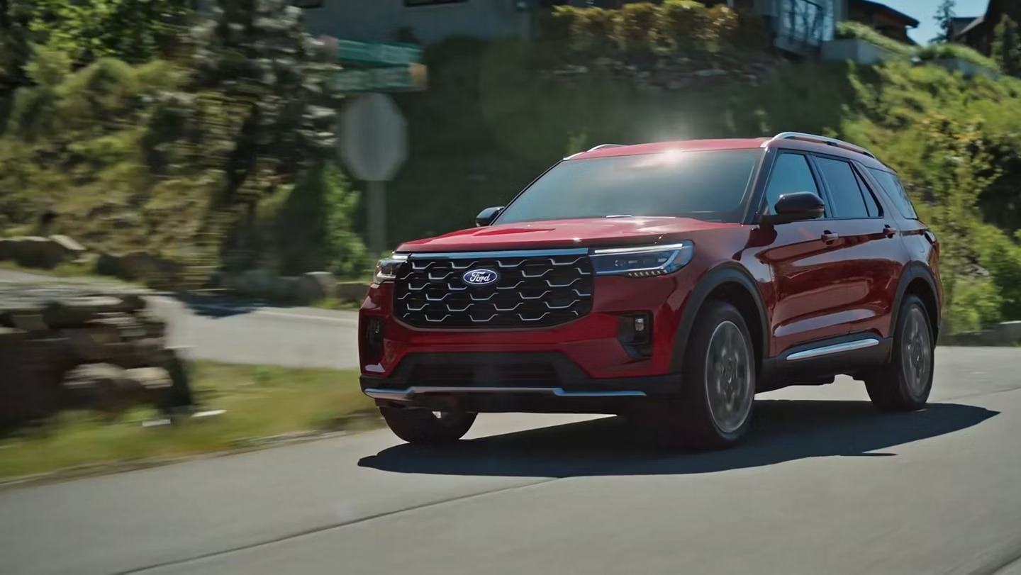 Red 2027 Ford Explorer SUV driving along a curving tree-lined road on a sunny day, shown from a front three-quarter angle with motion-blurred surroundings.