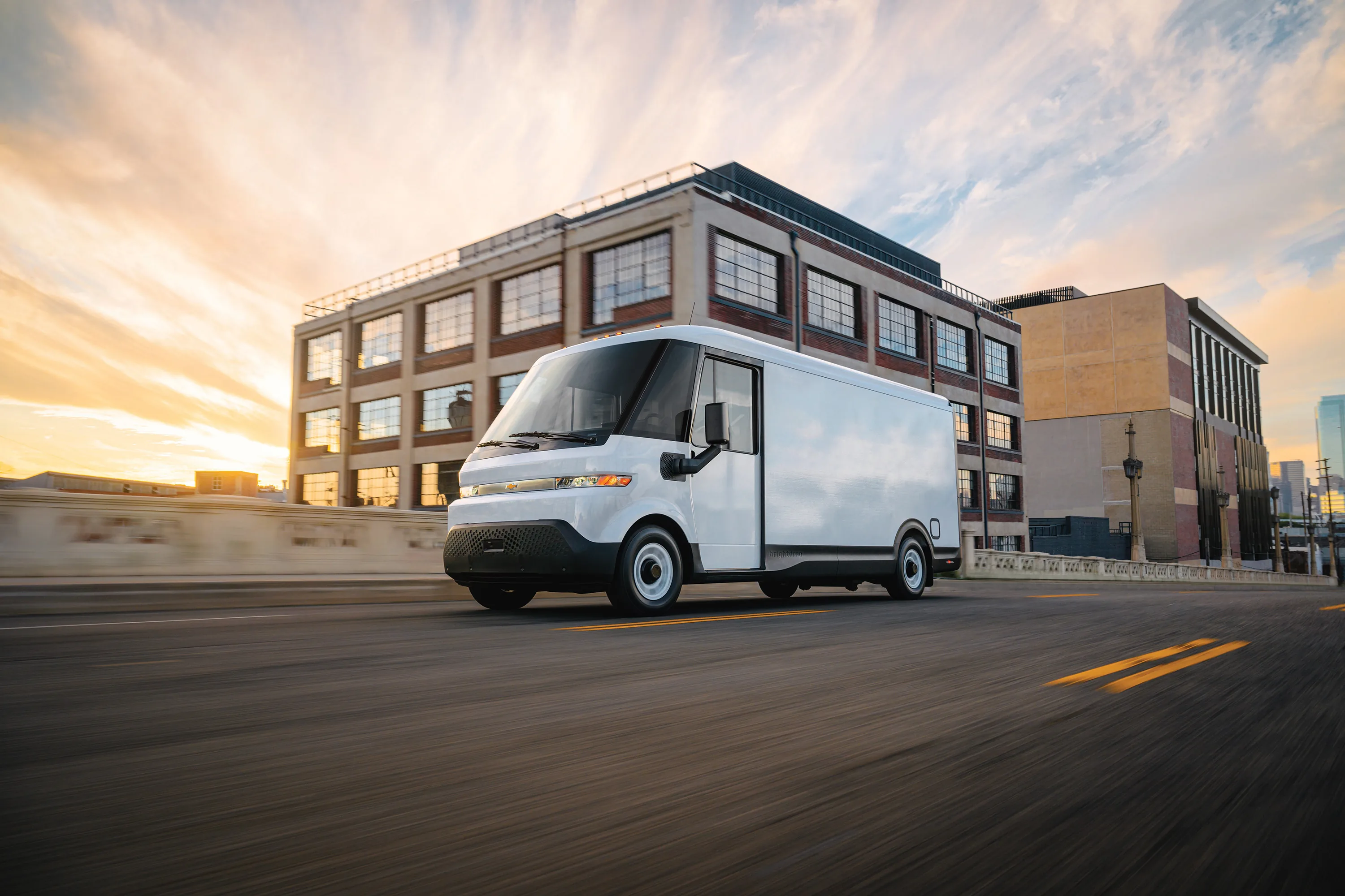 A white Chevrolet BrightDrop electric step van driving along a city street at sunset, with a legible Chevrolet bowtie badge on the grille and a brightdrop wordmark on the lower door panel, an industrial brick building in the background