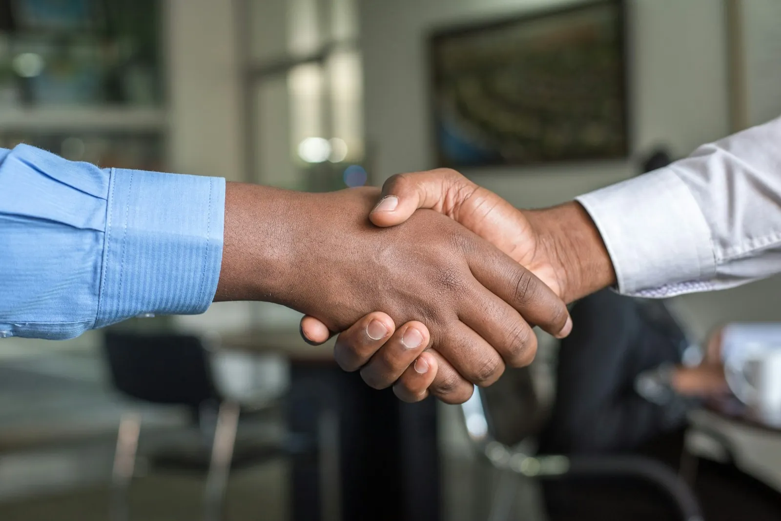 Close-up of two people in business attire shaking hands in an office, with a framed picture visible on the wall behind them.