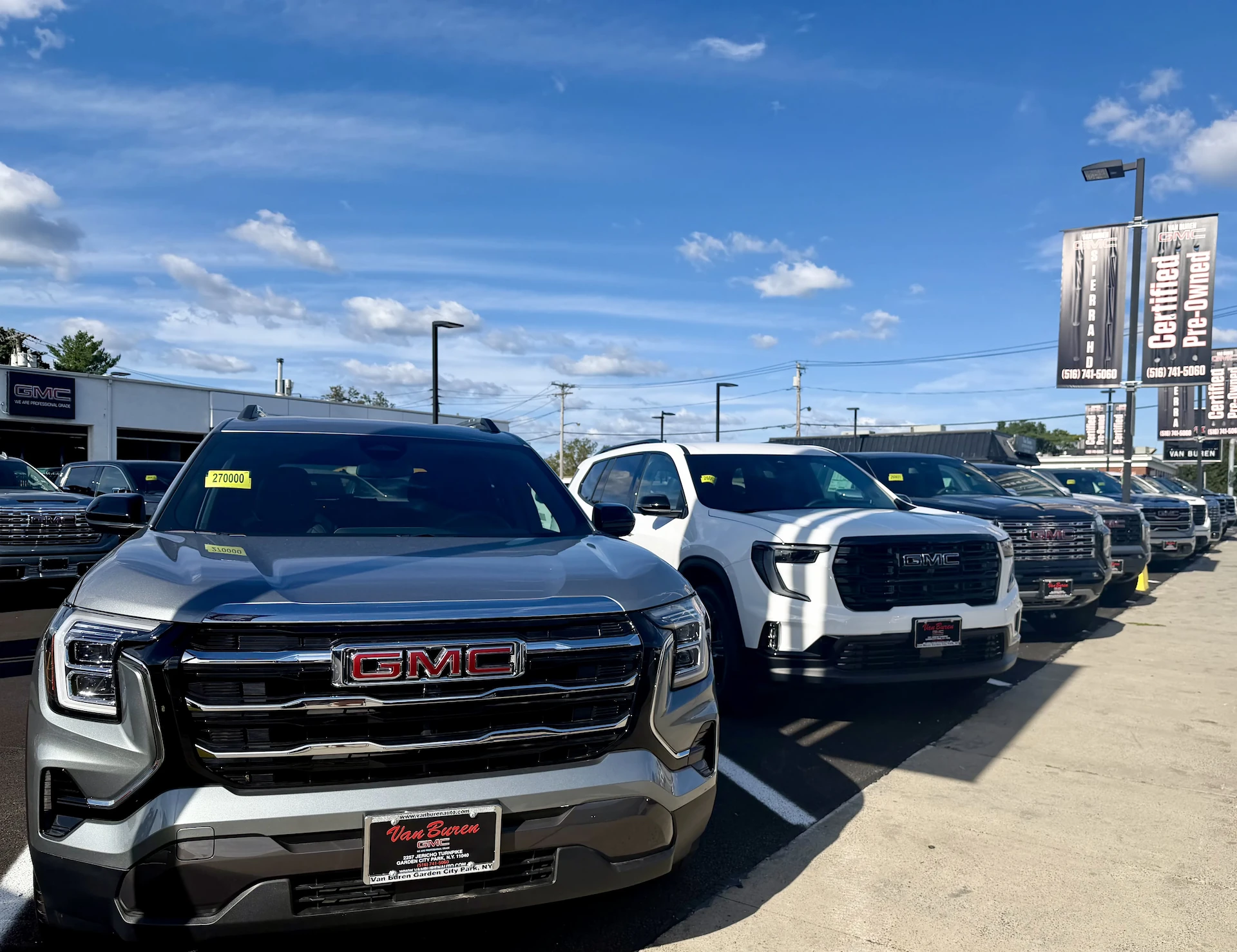 A row of GMC vehicles, led by a silver SUV and a white pickup truck each with a legible GMC grille badge, parked on the Van Buren GMC dealership lot; a Van Buren GMC dealer plate is visible on the nearest vehicle, with Certified Pre-Owned banners and the dealership building in the background under a blue sky