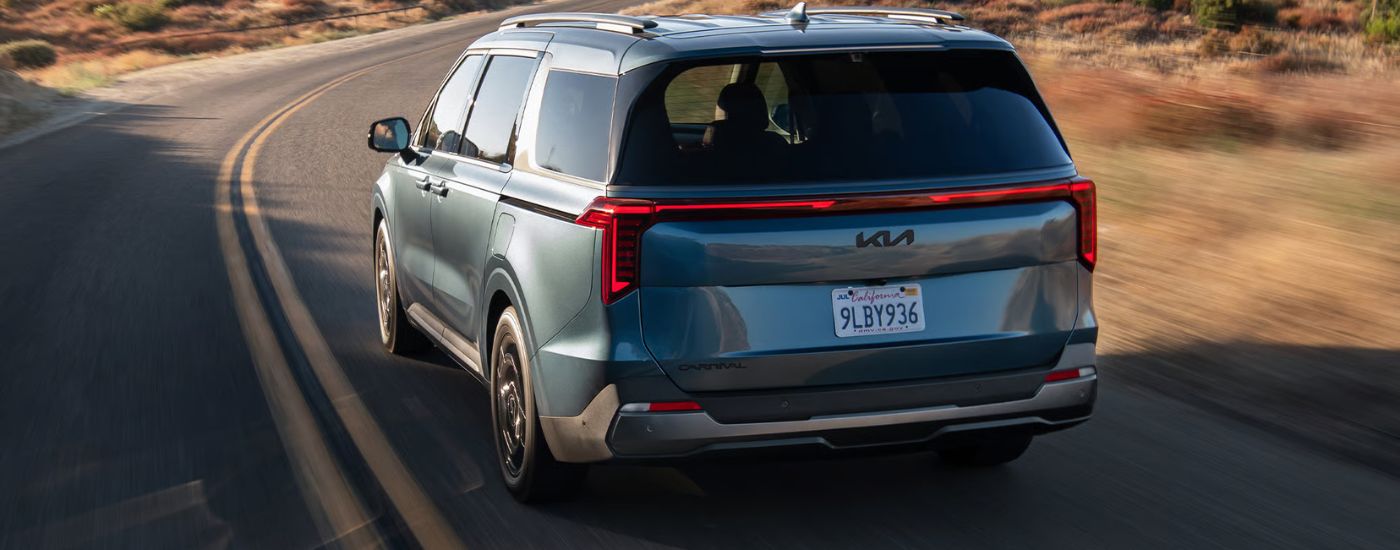 Rear view of a blue 2026 Kia Carnival driving on a paved road.