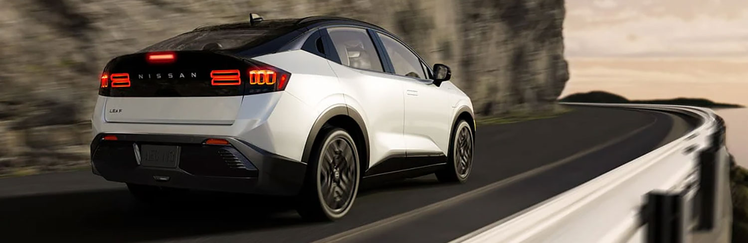 White 2026 Nissan Leaf electric crossover with a black roof driving along a winding coastal mountain road, shown from a rear three-quarter angle with motion-blurred surroundings.