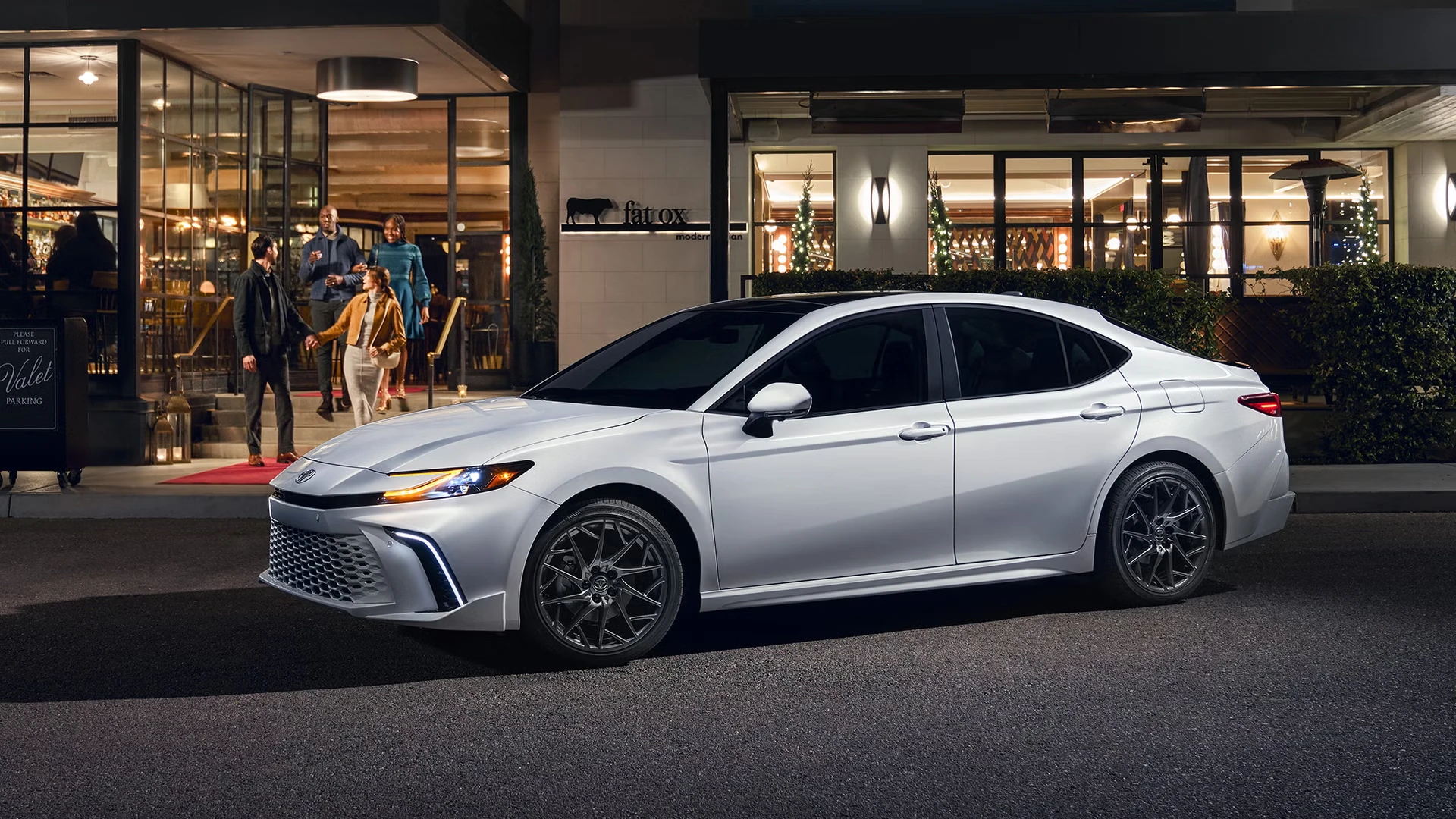 White Toyota Camry sedan with dark multi-spoke wheels and lit headlights, parked on a city street at night in front of a lit restaurant entrance with a group of people on the steps