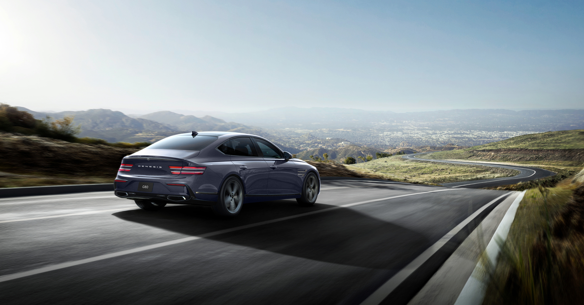 3/4 rear passenger side view of Genesis G80 Ceres Blue, in motion driving on road with rolling hills