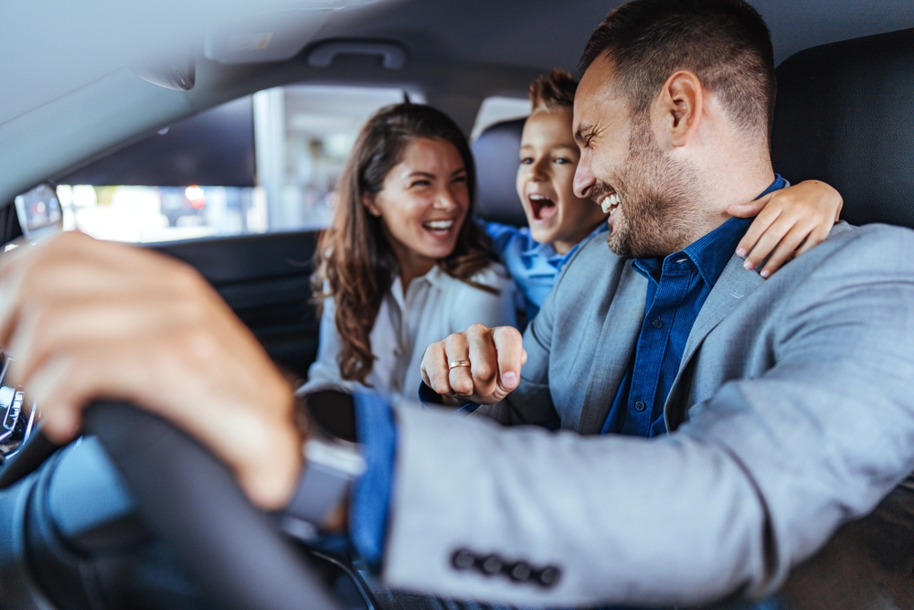 Happy family choosing new car, salesman showing them luxury auto at automobile dealership store. Customers selecting vehicle, consulting manager at modern showroom shop