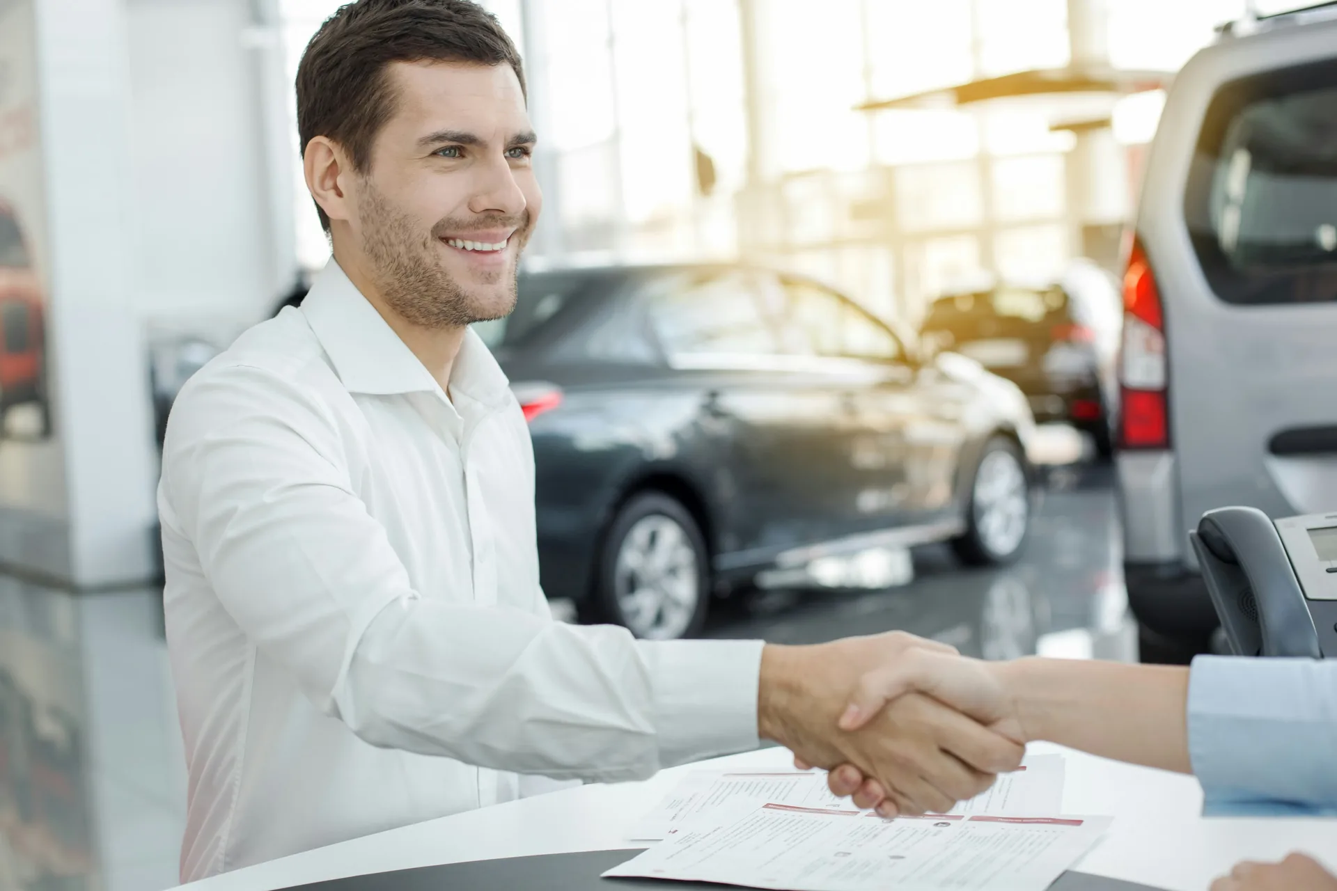 Salesperson and customer shaking hands over a purchase agreement at a dealership desk, with vehicles visible in the showroom behind them