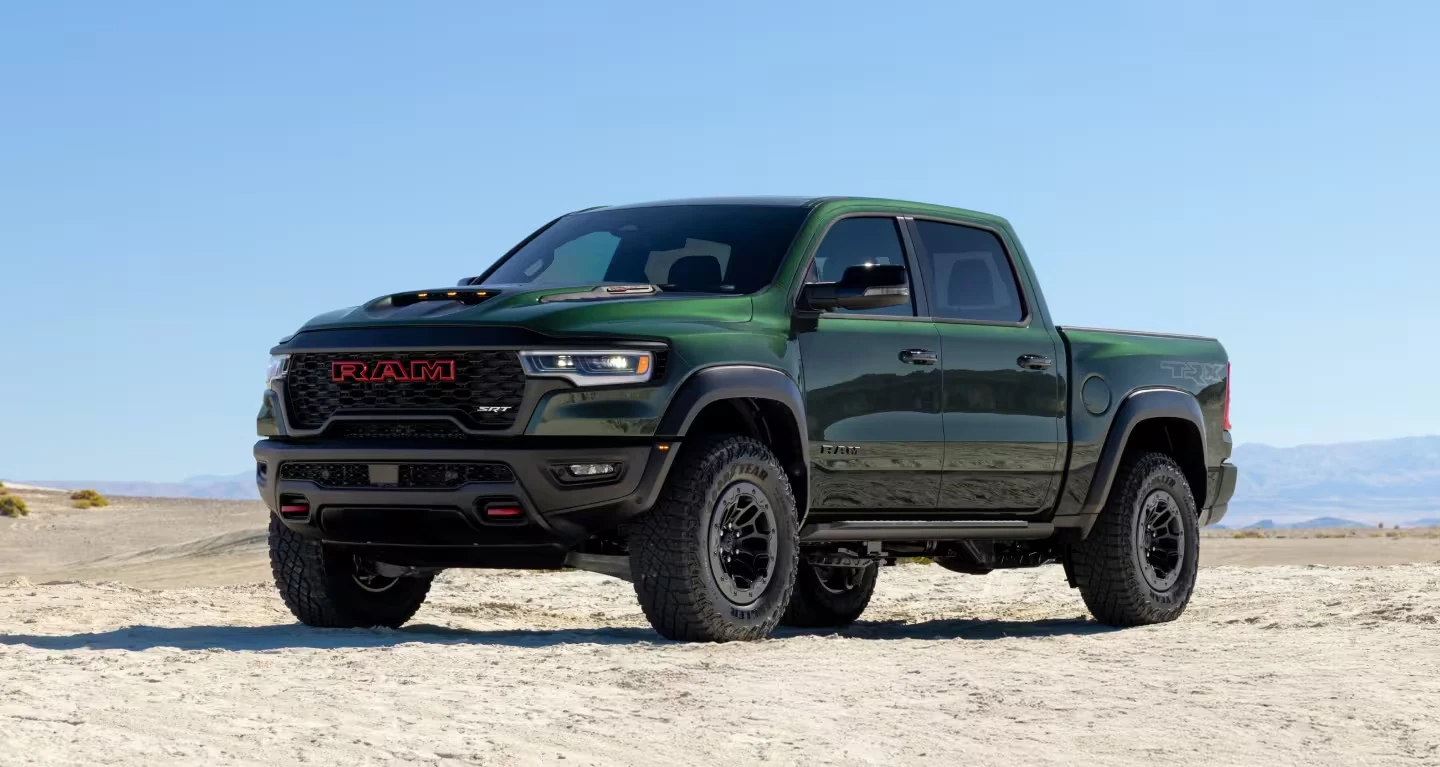 Dark green 2027 Ram TRX crew cab pickup with a red-outlined RAM grille, hood scoop, black wheels and large off-road tires, parked on pale desert ground under a clear blue sky with distant mountains