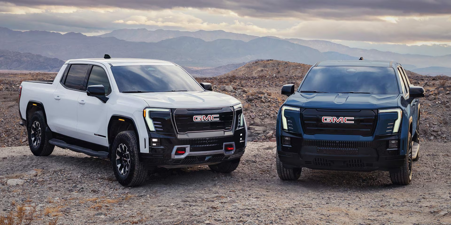 White GMC Sierra EV AT4 and blue GMC Sierra EV parked side by side in the desert