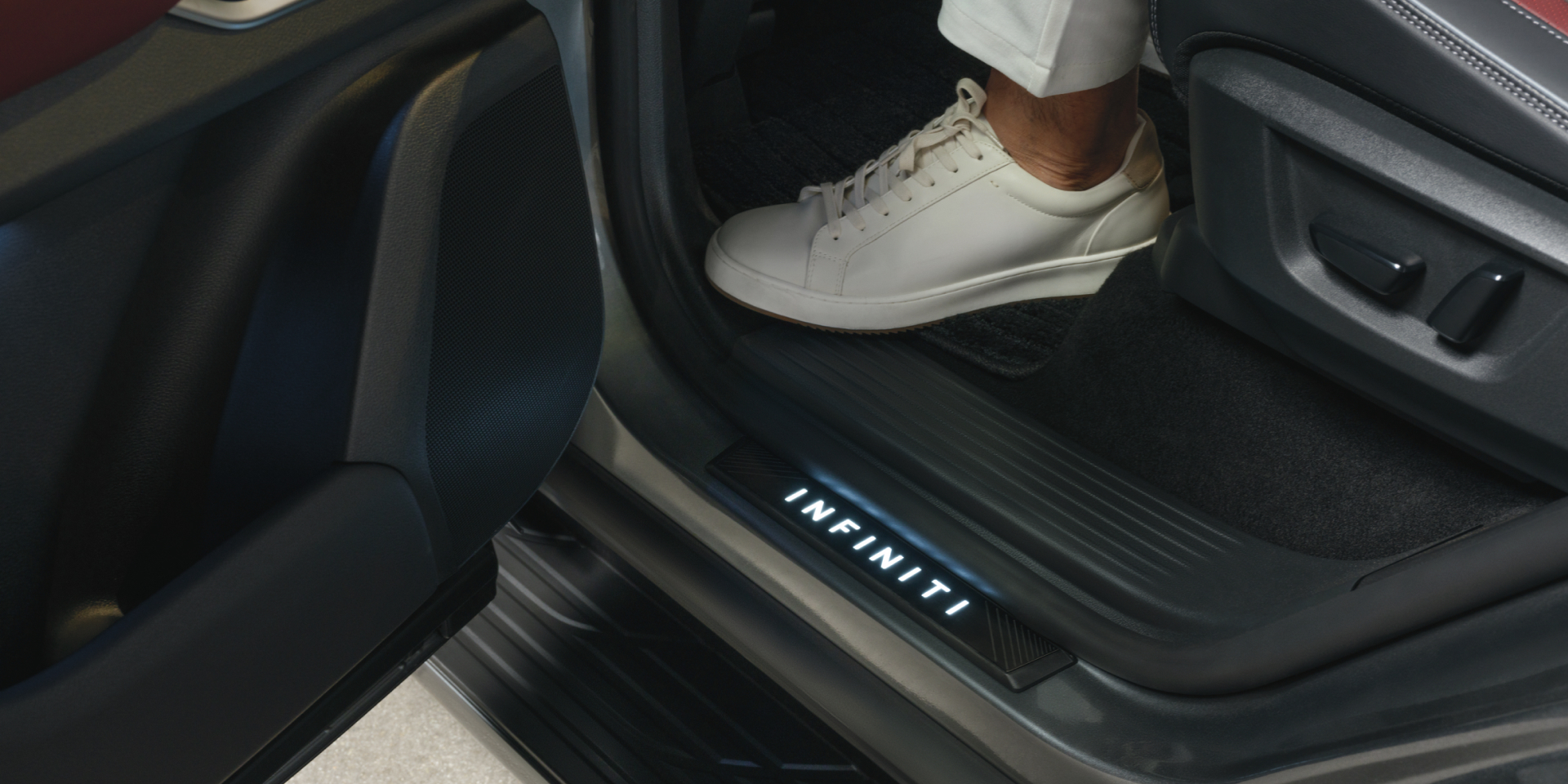 Close-up of the illuminated INFINITI kick plate on the 2027 INFINITI QX80 doorsill as a passenger steps into the cabin.
