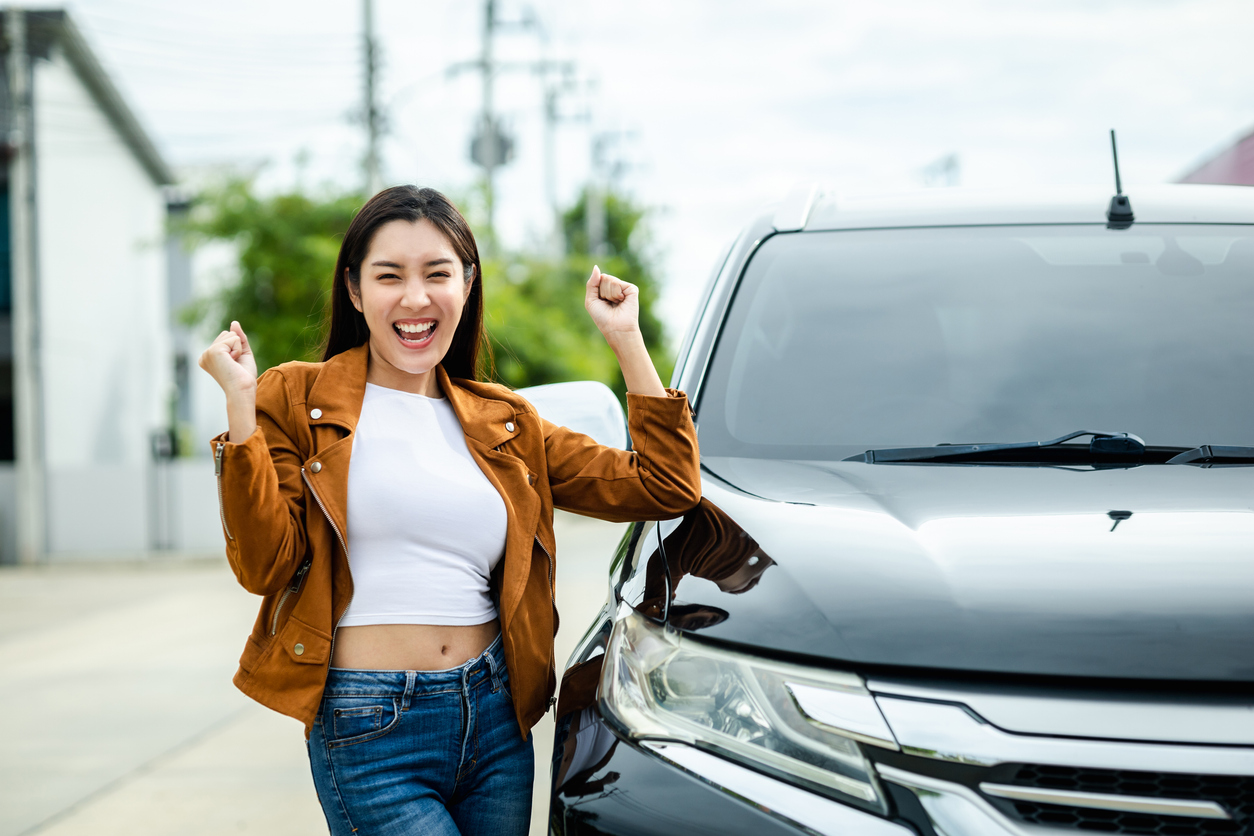 Happy woman smiling while standing beside her parked car in a sunny outdoor setting.
