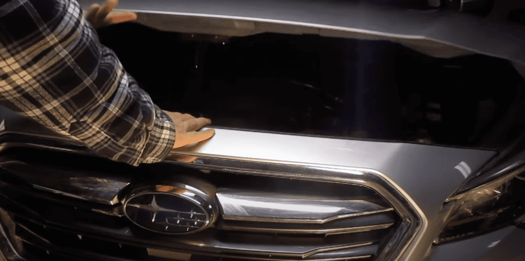 Hand reaching for the hood latch above a Subaru grille