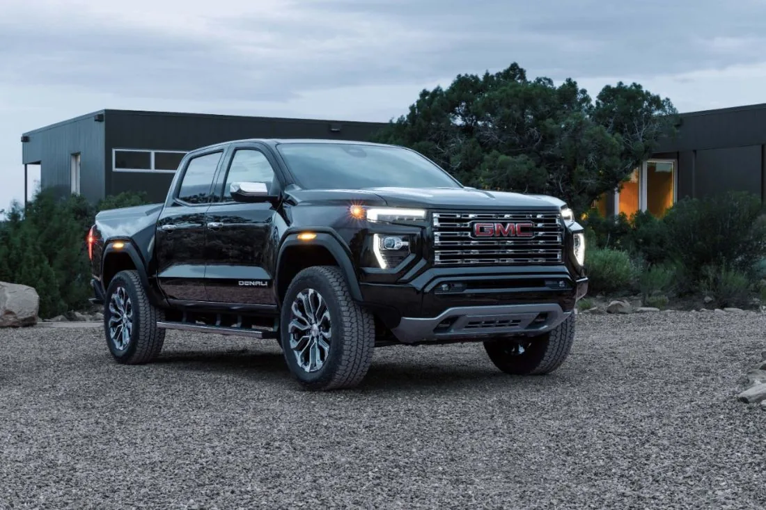 Front three-quarter view of a black GMC pickup truck with a Denali badge, parked in front of a modern house at dusk with its headlights on