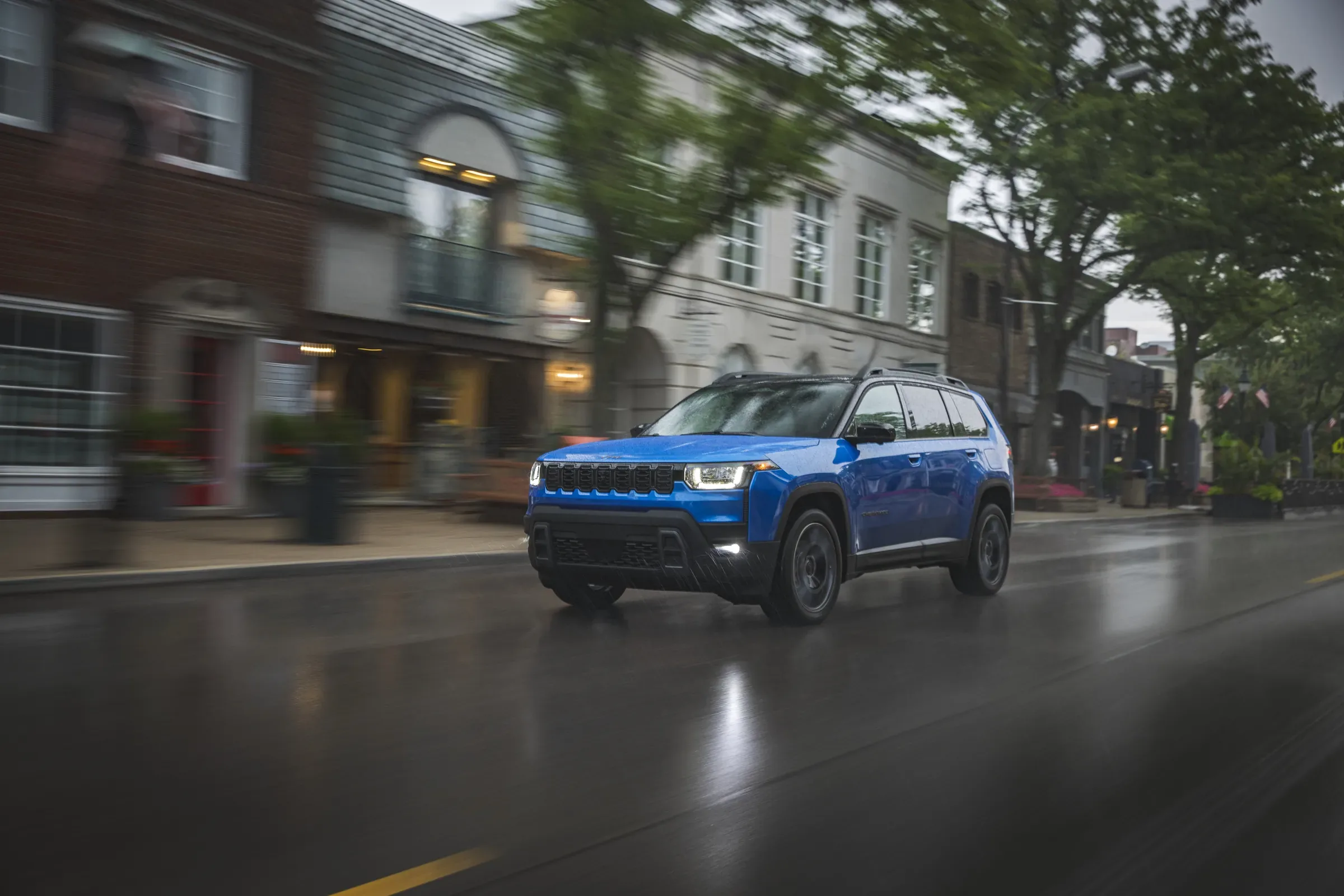 Blue Jeep Cherokee driving through a rain-slicked city street lined with historic buildings and trees
