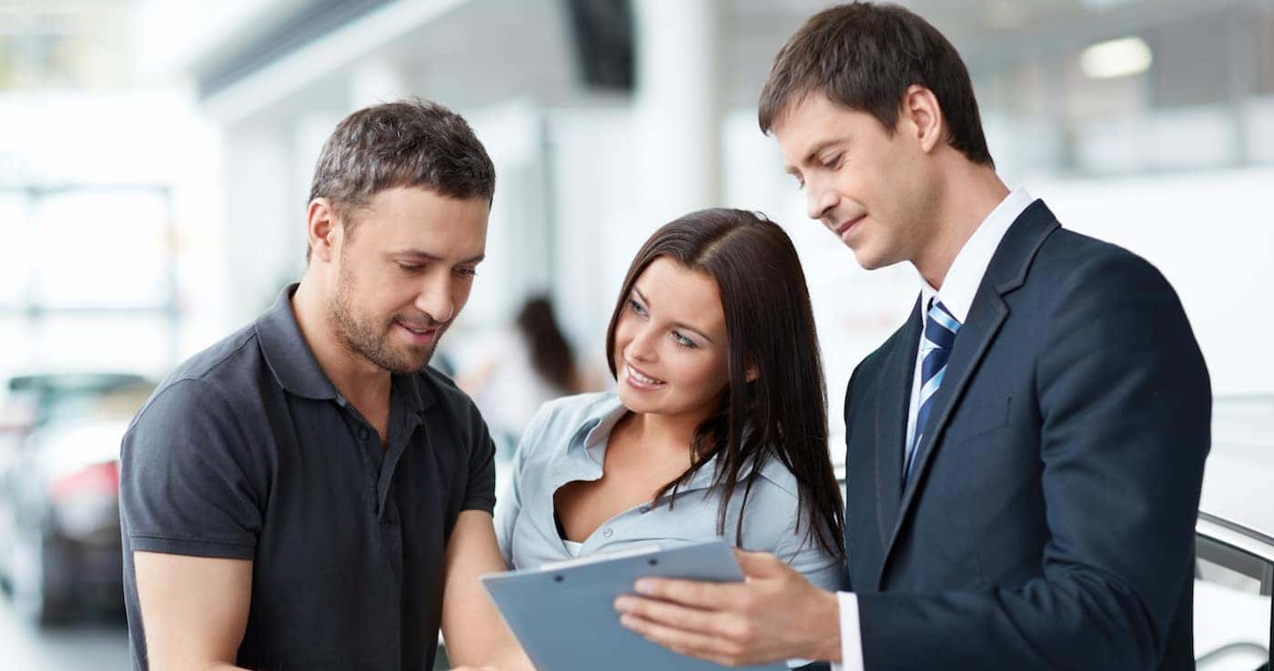 Hero image of a car salesman reviewing an auto loan with a young couple.