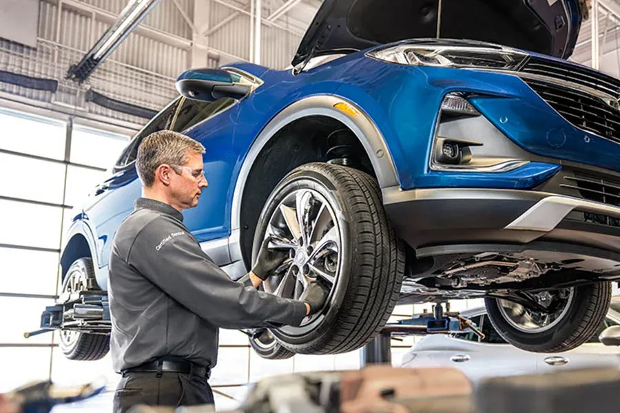 Technician wearing a "Certified Service" branded uniform works on a wheel of a blue SUV raised on a lift, with the hood open