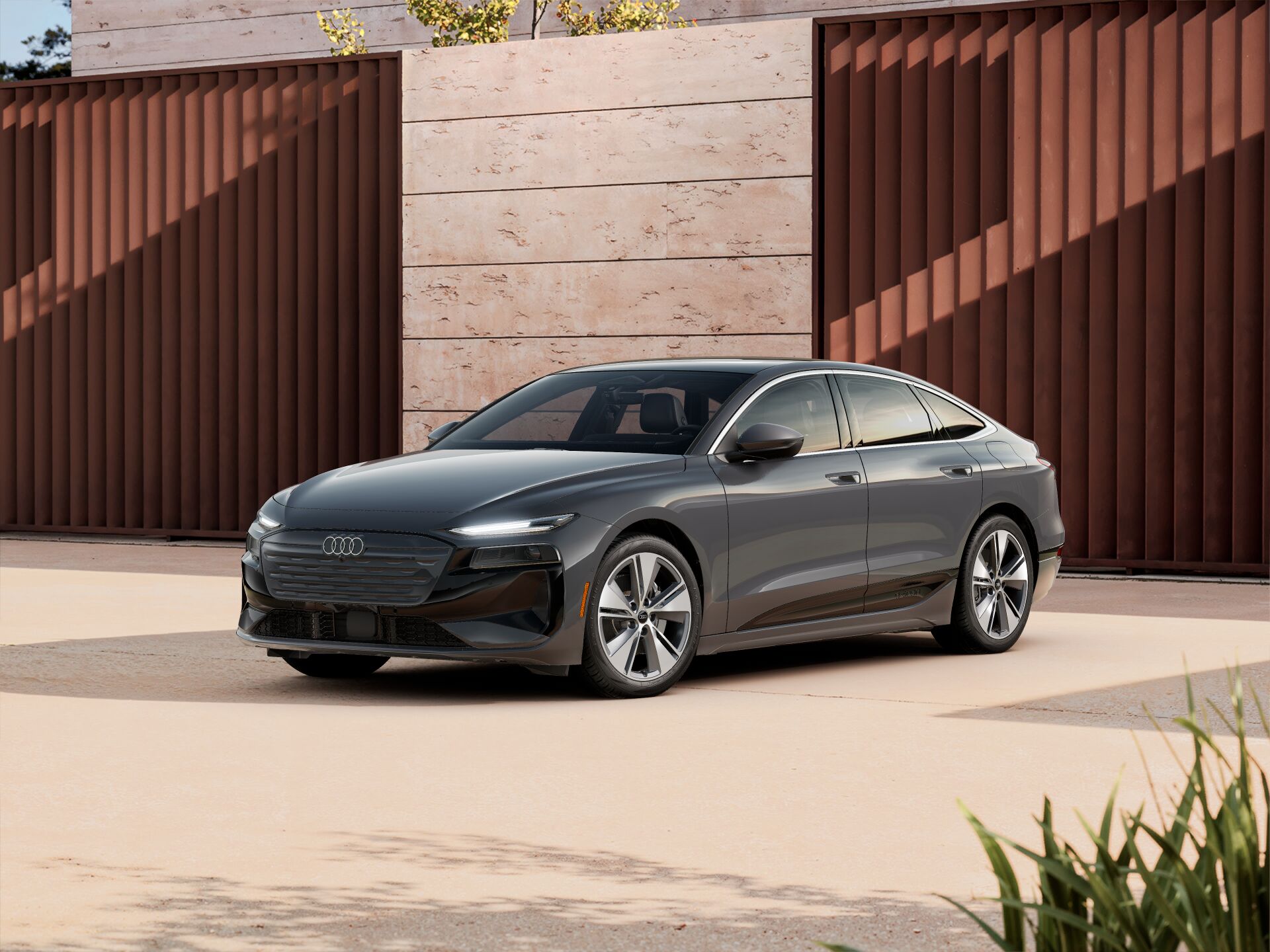 Dark gray 2027 Audi A6 Sportback e-tron electric sedan parked on a paved area in front of a modern stone and metal wall, shown from a front three-quarter angle.
