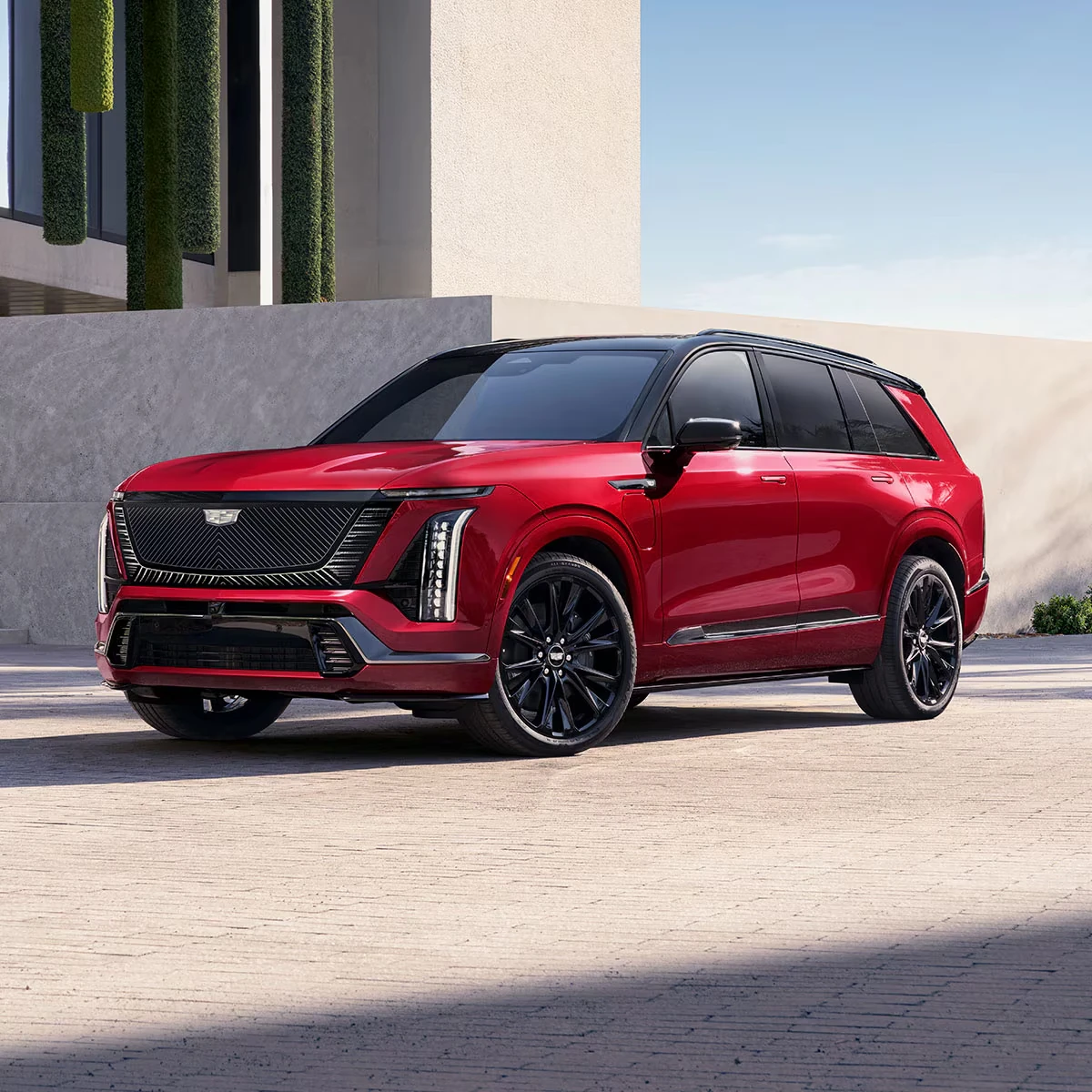 Red 2027 Cadillac Vistiq three-row SUV with a black roof, black multi-spoke wheels, mesh grille and vertical LED headlights, parked on a brick plaza beside a concrete wall and modern building