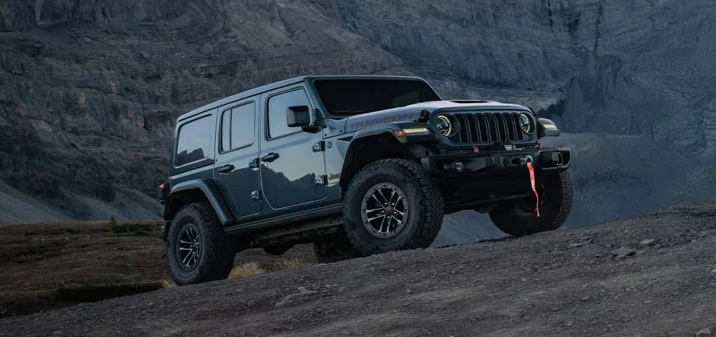 Dark gray 2026 Jeep Wrangler Rubicon four-door SUV with a front winch on a rocky slope, with a steep rock wall behind it