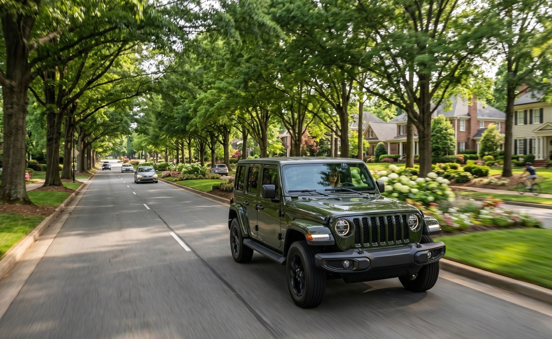A green Jeep Wrangler SUV driving down a tree-lined residential suburban street on a sunny day.