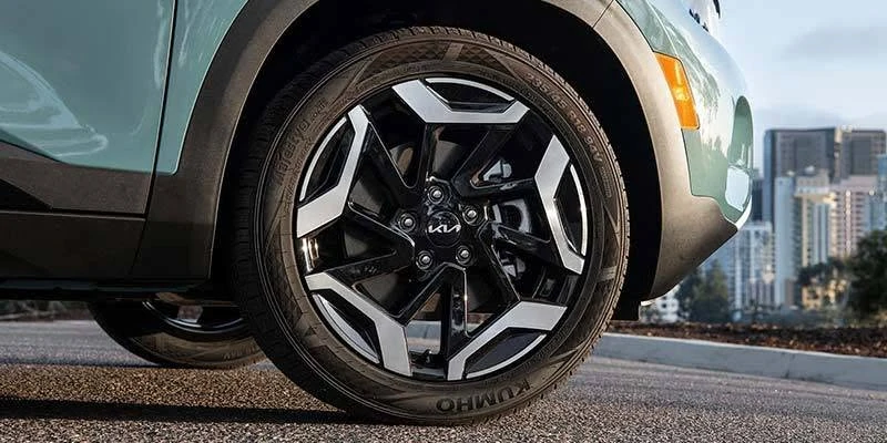 Close-up of a Kia SUV’s black machined-face alloy wheel with a Kia-branded center cap and Kumho tire, set against a city skyline backdrop.