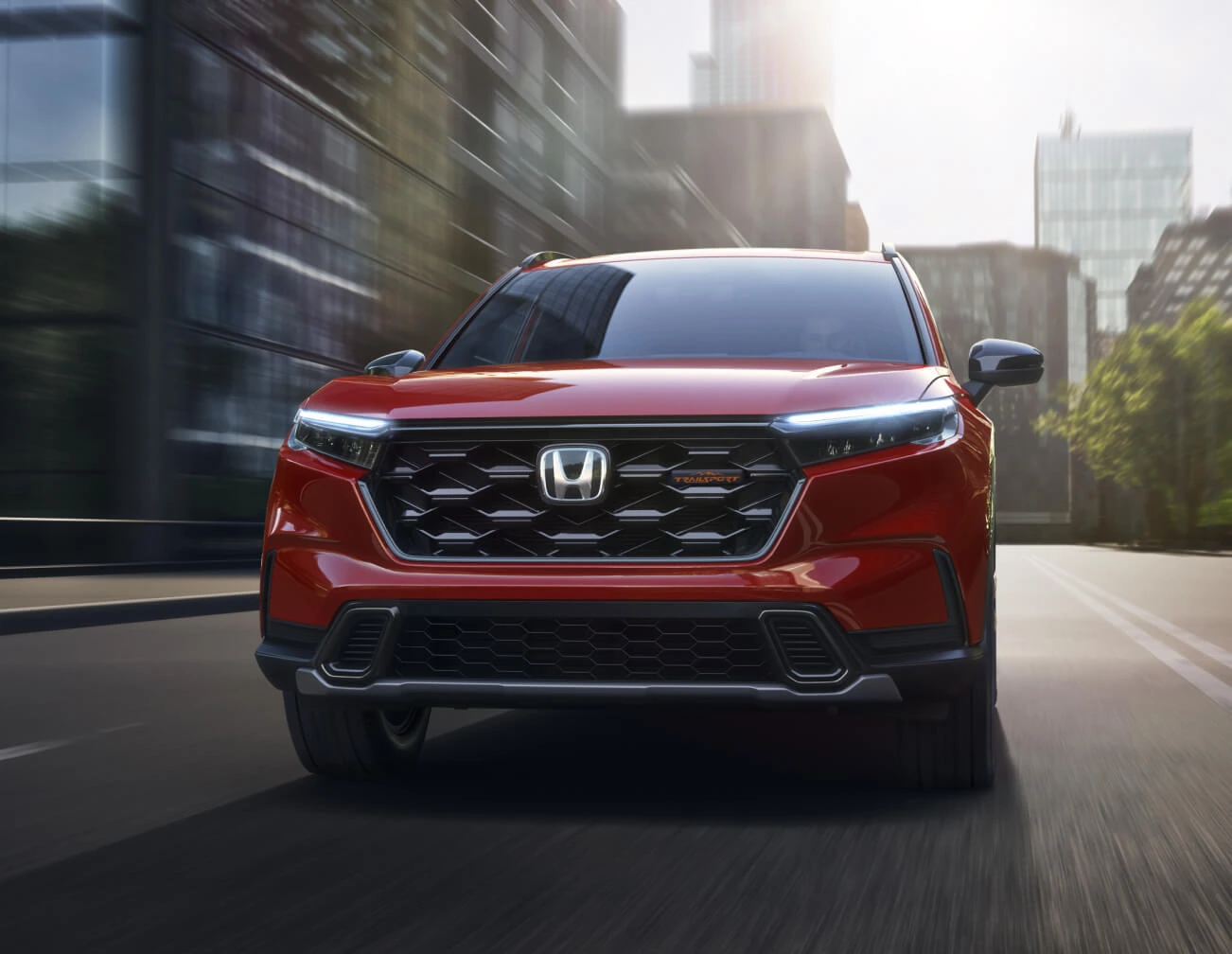 2026 Honda CR-V TrailSport Hybrid in Radiant Red Metallic, front view driving through a city street at sunrise.