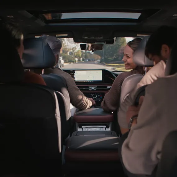 Interior view from the middle row of a 2027 INFINITI QX80 with four occupants, a wide navigation display, burgundy console accents, and a panoramic sunroof open to bright sky.