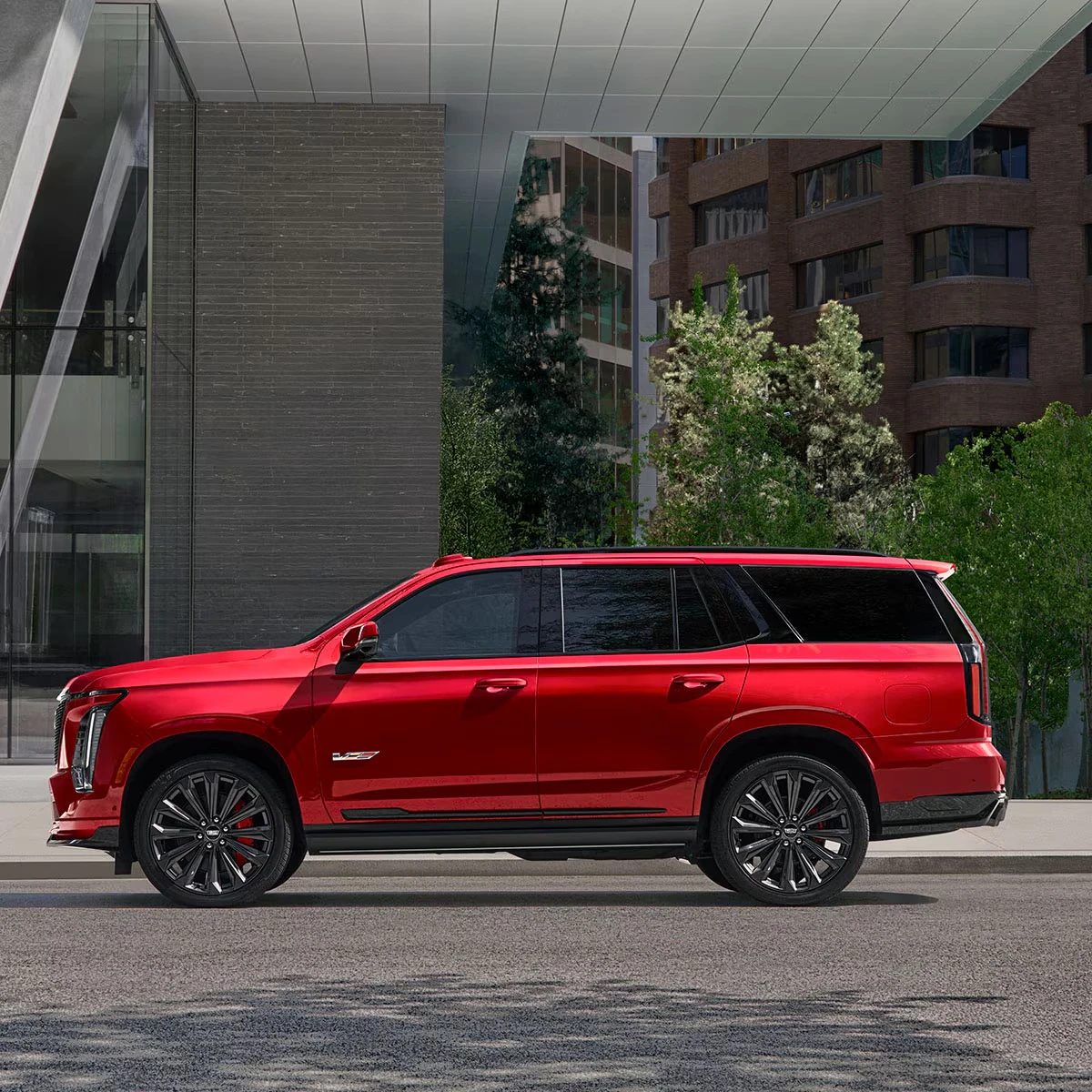 Red 2027 Cadillac Escalade-V SUV in side profile with black multi-spoke wheels, red brake calipers and a black roof line, parked on a city street in front of glass and brick buildings with green trees