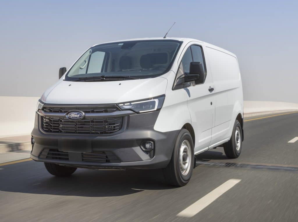 White Ford Transit Custom cargo van driving on a highway, shown from a front three-quarter angle.