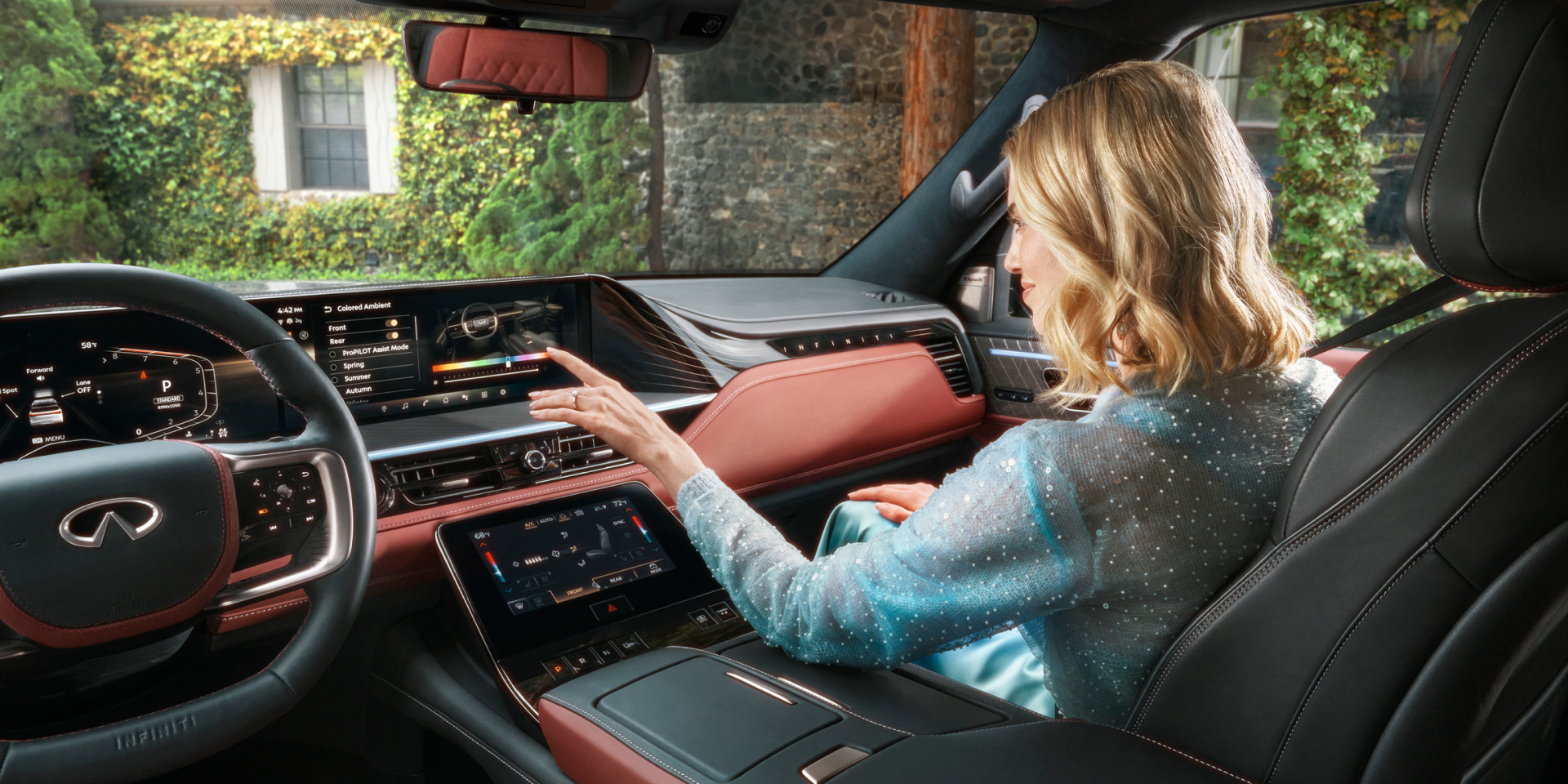 A passenger in the 2027 INFINITI QX80 touches the wide dual-screen infotainment display while adjusting colored ambient lighting in a burgundy and black leather cabin.