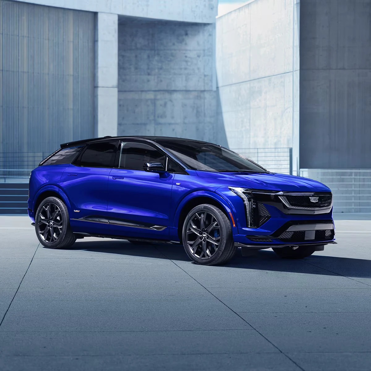 Blue Cadillac Optiq-V SUV with a black roof, black multi-spoke wheels, blue brake calipers and a mesh grille, parked on a concrete plaza between gray architectural walls