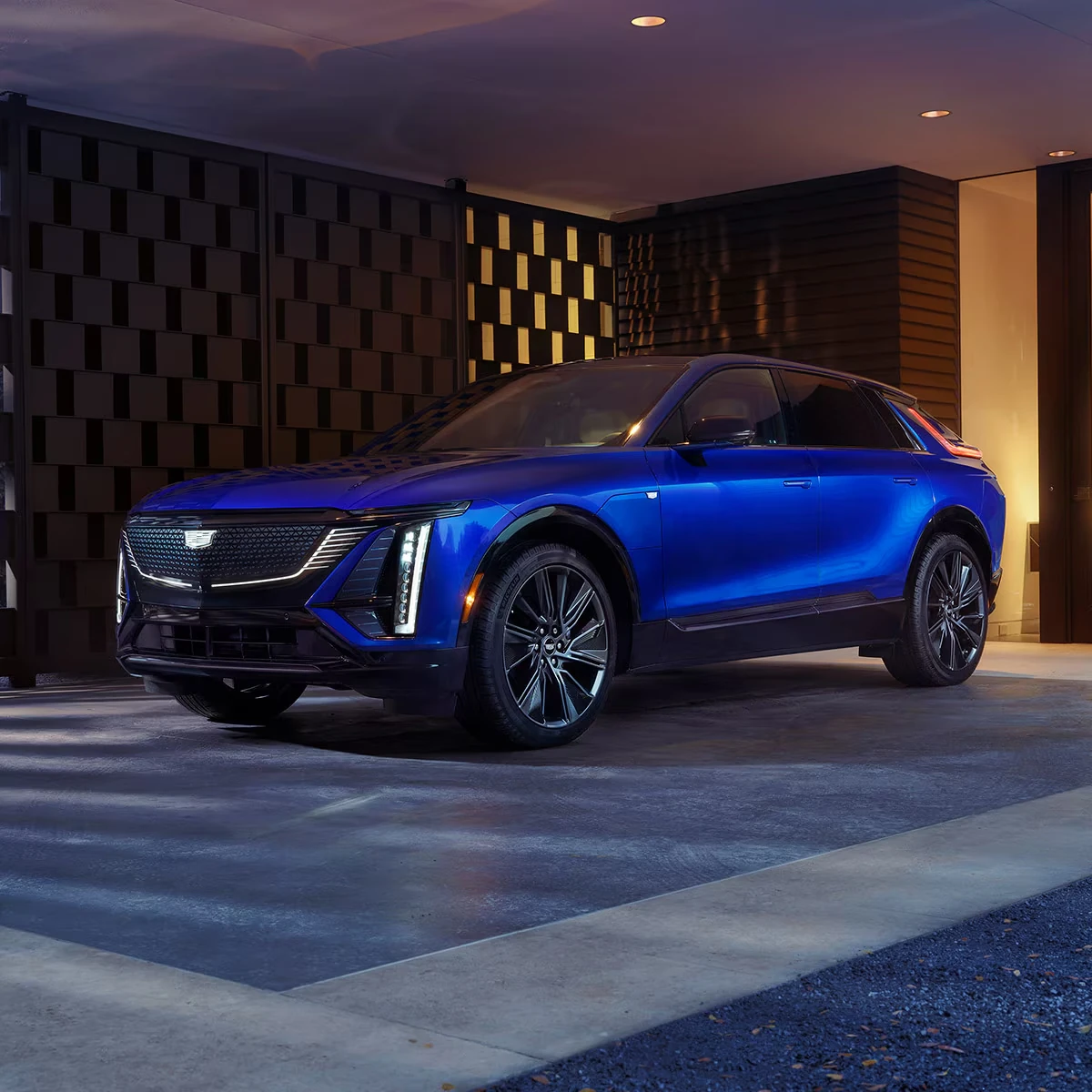 Bright blue 2027 Cadillac Lyriq SUV with illuminated vertical headlights and dark multi-spoke wheels, parked under a covered entry beside a patterned screen wall in evening light