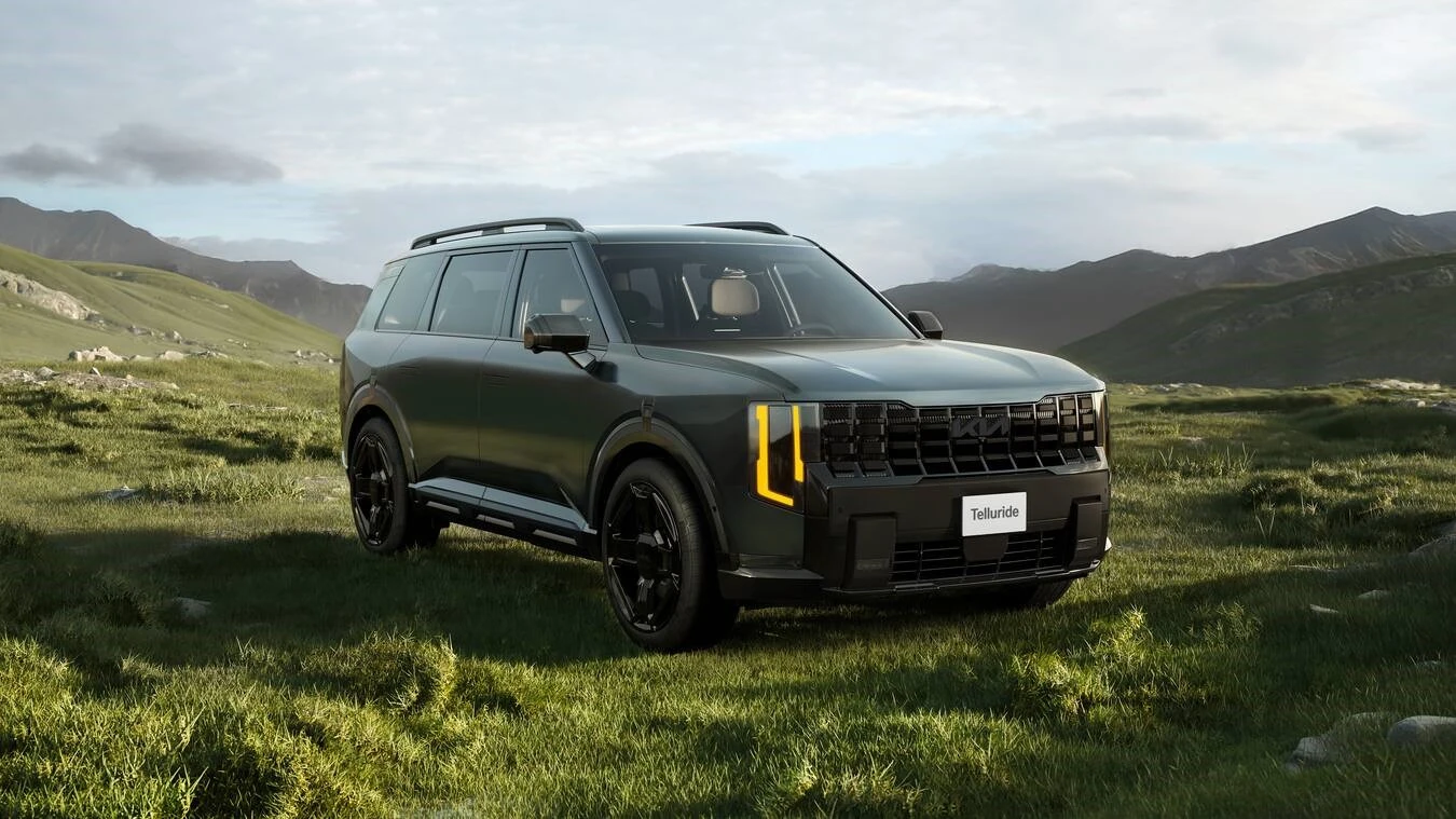 Matte dark green 2027 Kia Telluride HEV X-Line, front three-quarter view, parked on a grassy hillside with rolling green mountains behind it and amber-accented headlights illuminated.