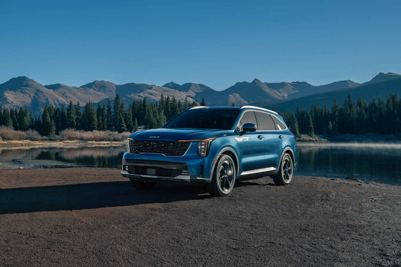 Blue 2026 Kia Sorento Hybrid parked at a mountain lake overlook, with pine forest and mountain peaks reflected in the still water.