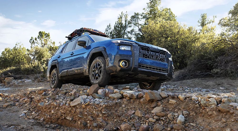 A blue 2027 Subaru Outback Wilderness is driving off-road on rocky terrain.