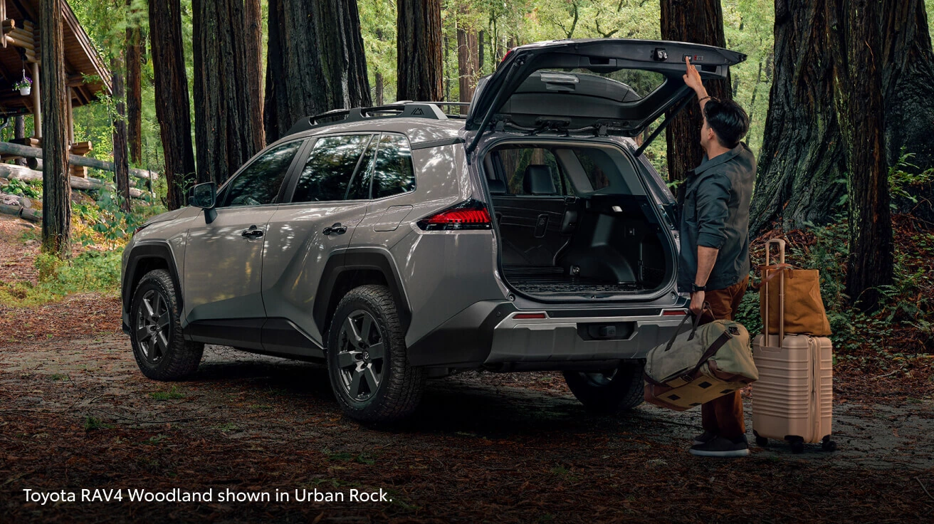Side view of a man pressing the tailgate button on a 2026 Toyota RAV4 Woodland Edition in Urban Rock in a forest.